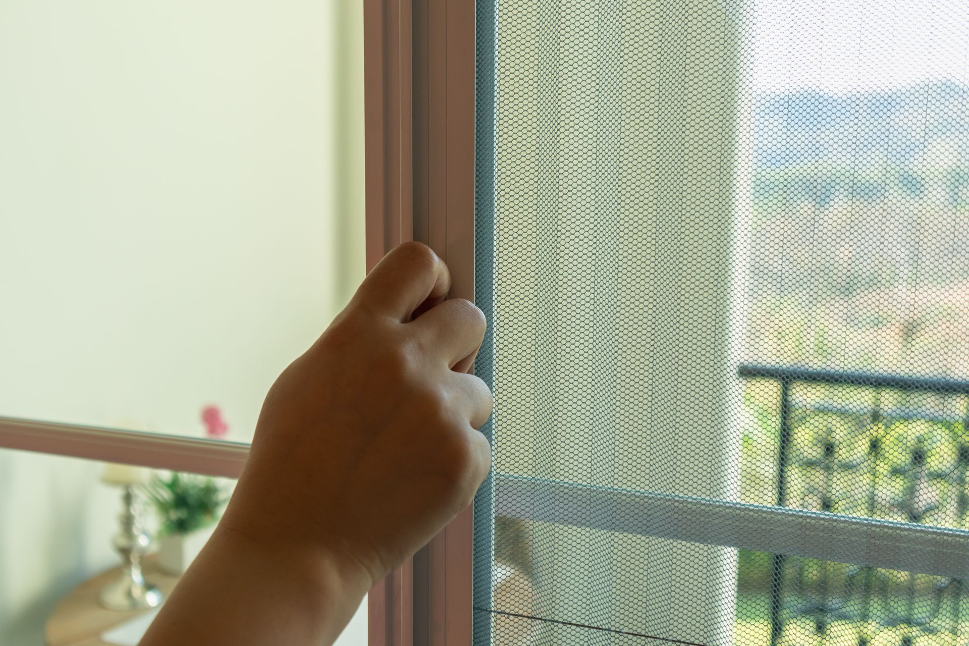 Hand pulling back a screen door to reveal a sunlit balcony and greenery outside