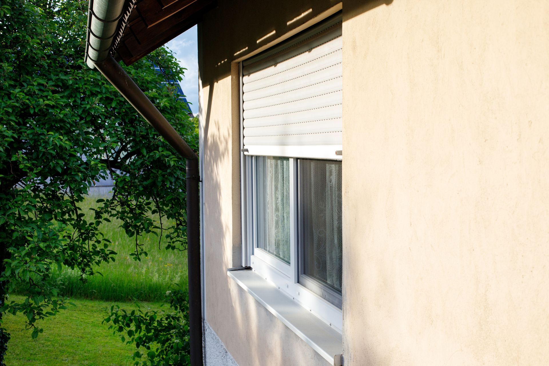White house wall with window beside a green garden and tree, viewed from under the roof eaves