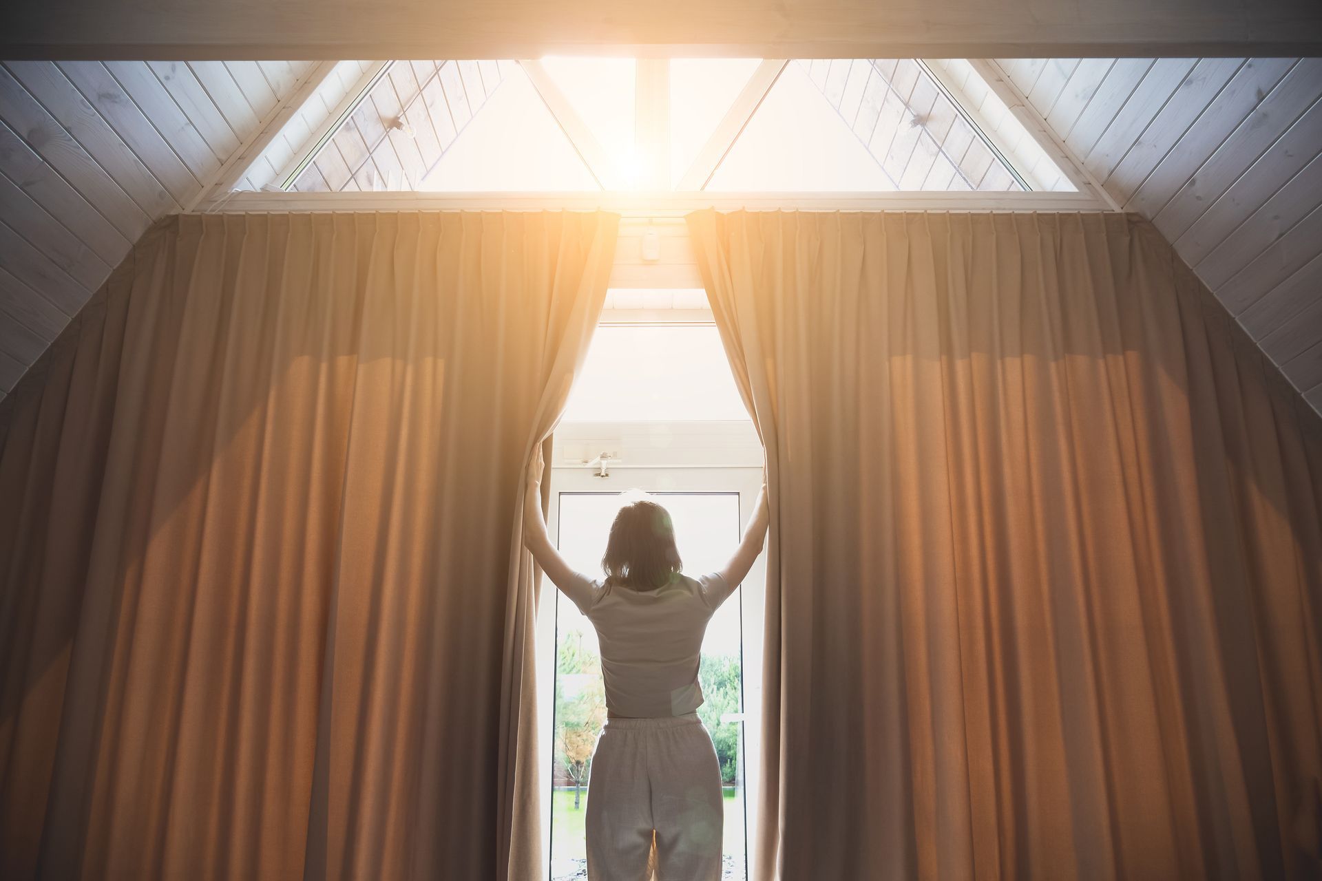 Person standing by a bright window with arms raised, framed by tall curtains in a softly lit room