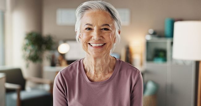Smiling woman in a mauve shirt seated indoors, with a softly blurred home background