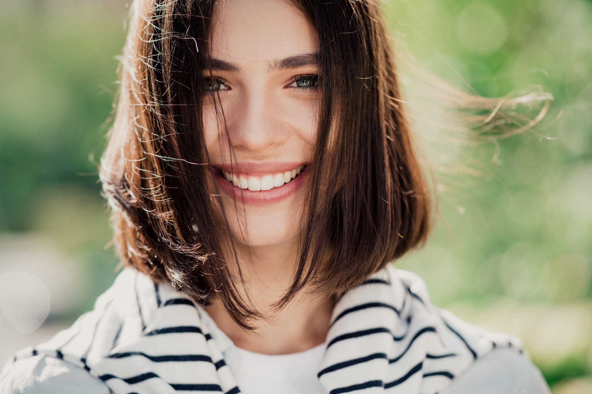 Smiling person with short brown hair in a striped shirt outdoors with a blurred green background