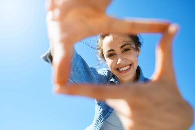 Smiling person framed through fingers against a bright blue sky