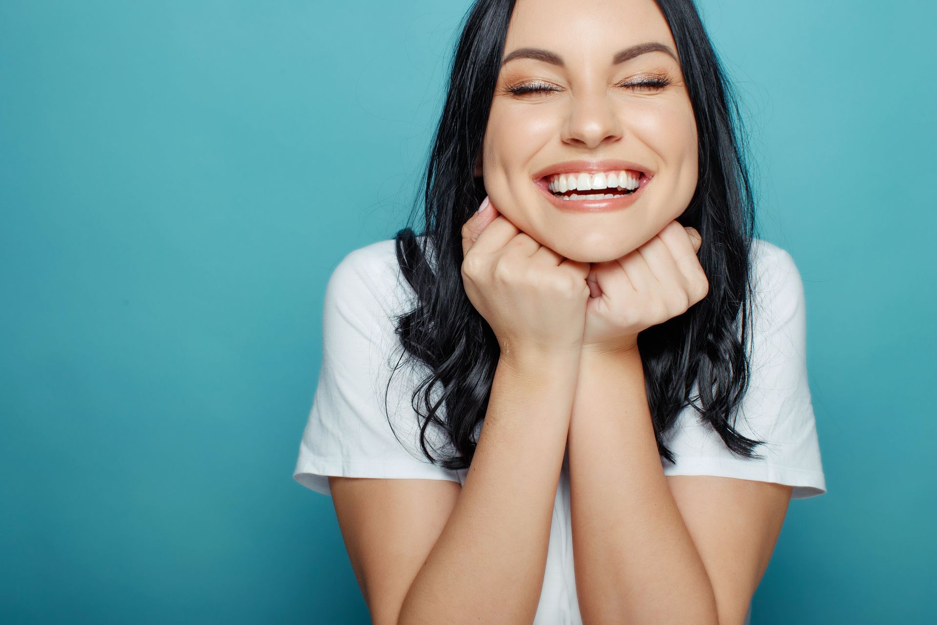 Smiling woman with dark hair resting her chin on her hands against a teal background