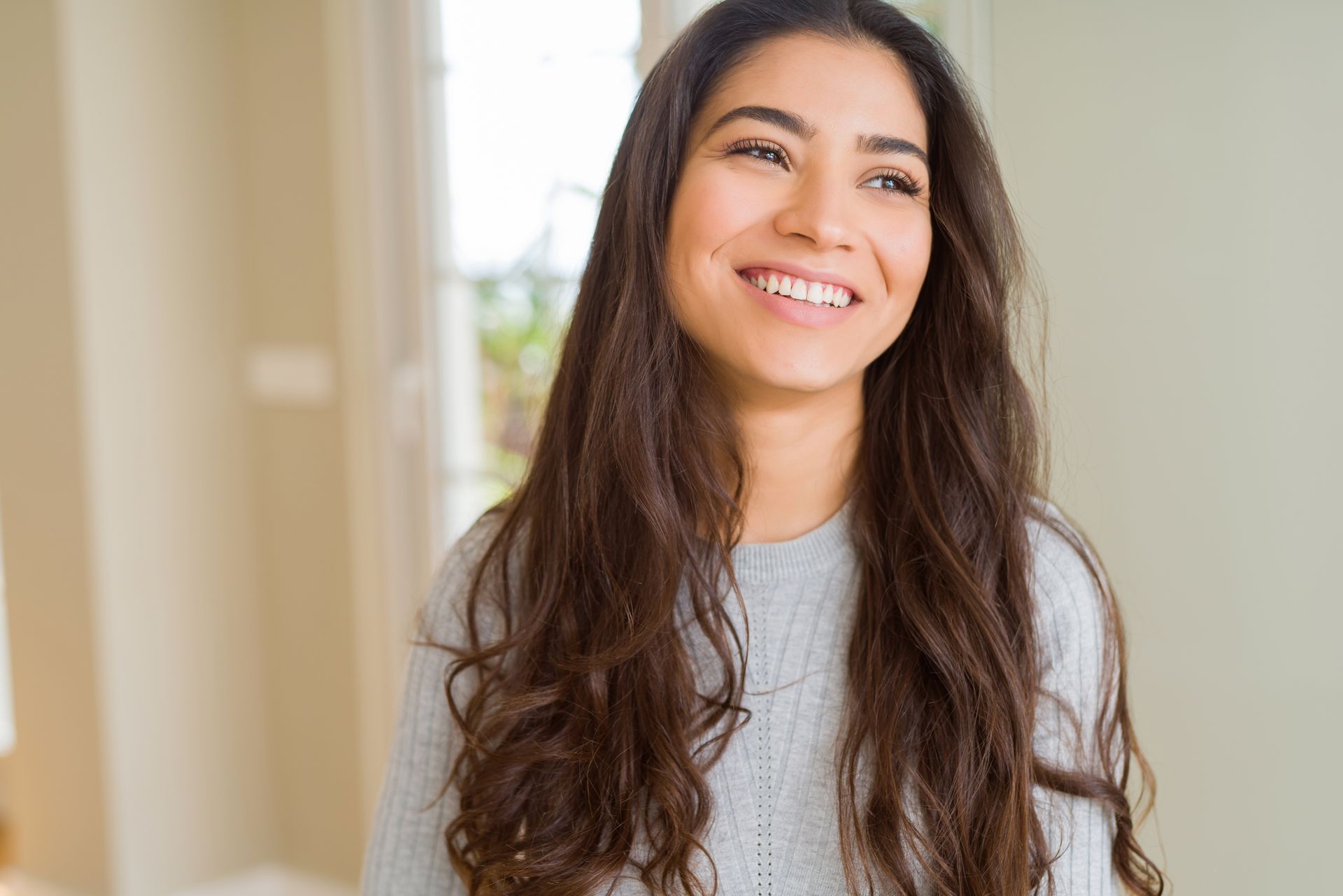 Smiling woman with long wavy hair in a light gray sweater, standing indoors near a bright window.