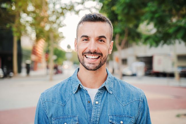 Smiling man in a denim shirt standing on a tree-lined city street