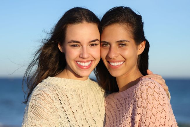 Two smiling women standing close together outdoors against a blue sky, wearing cream and pink sweaters