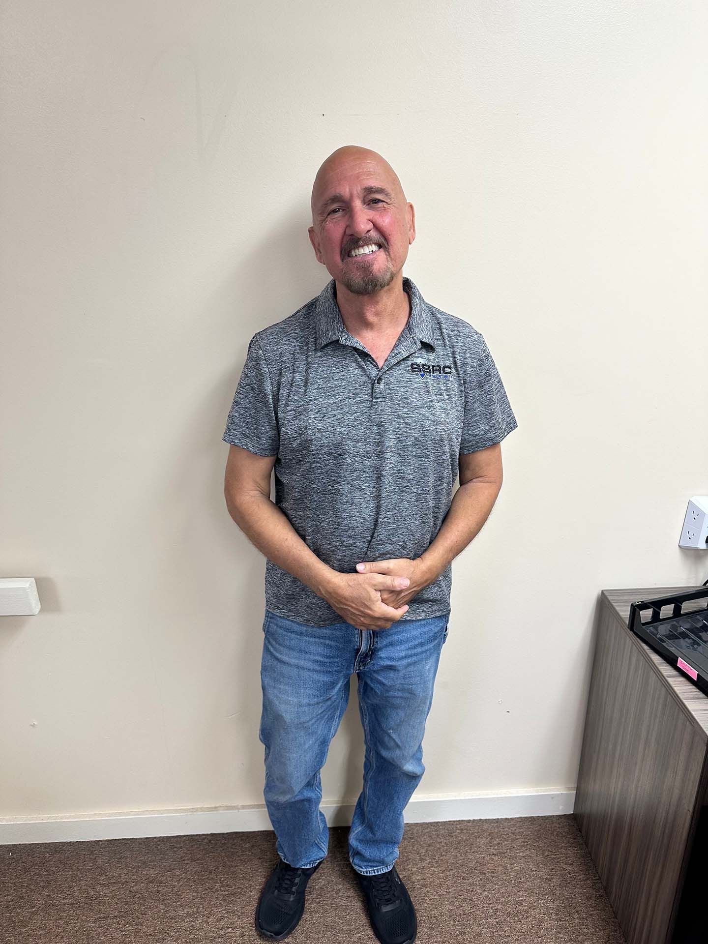 Smiling man in patterned shirt and jeans standing against a beige wall, hands clasped in front