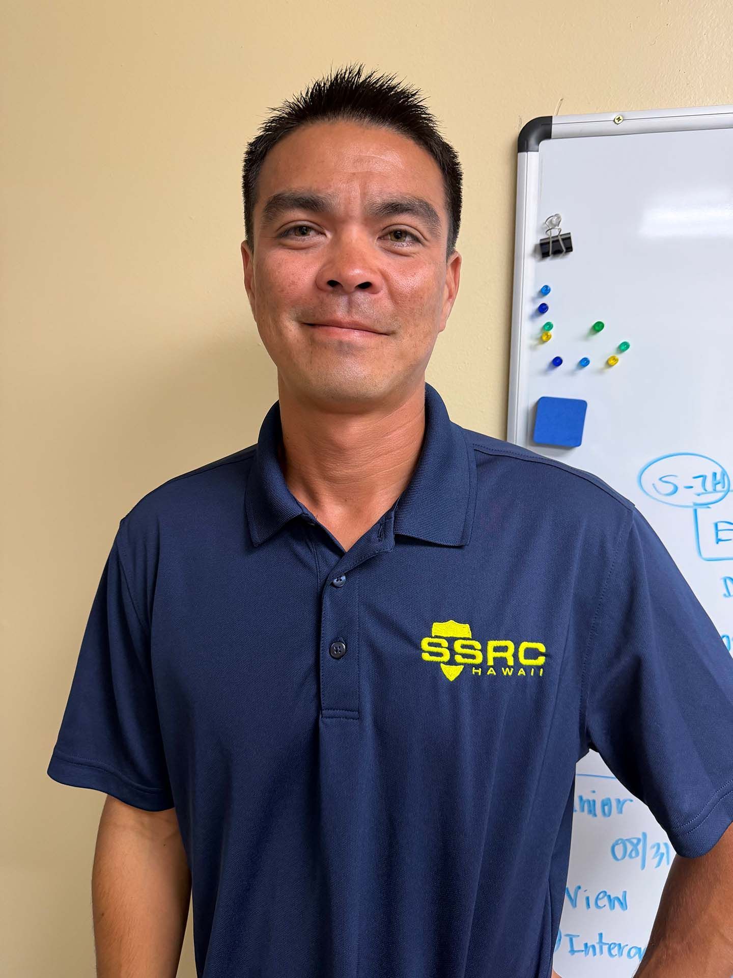 Man in navy polo standing beside a whiteboard in a classroom or office, smiling at the camera