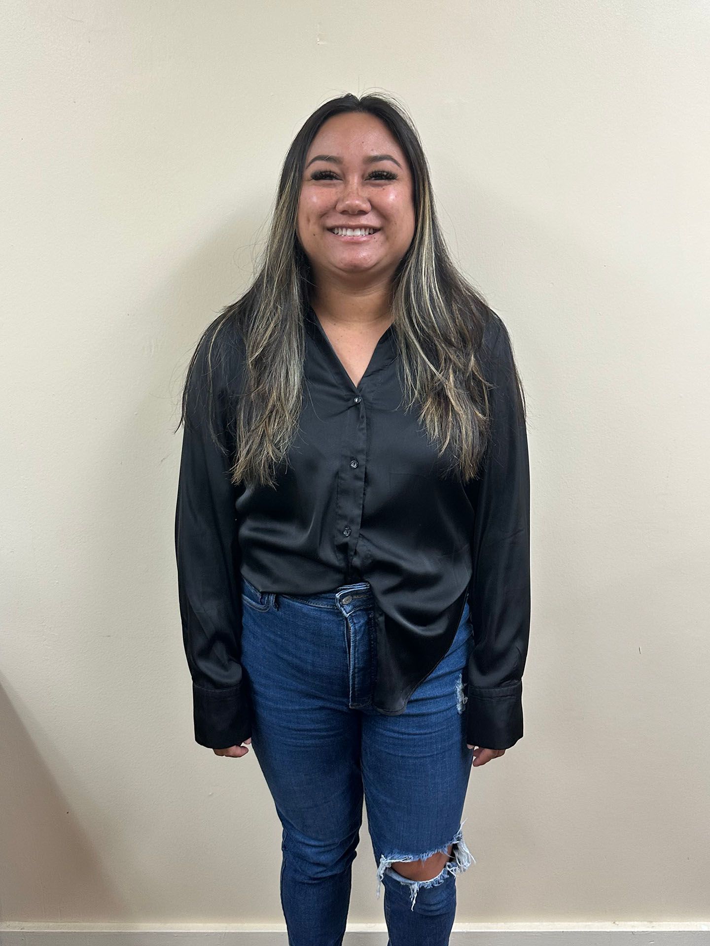 Standing against a beige wall, smiling person wears a black blouse and blue jeans