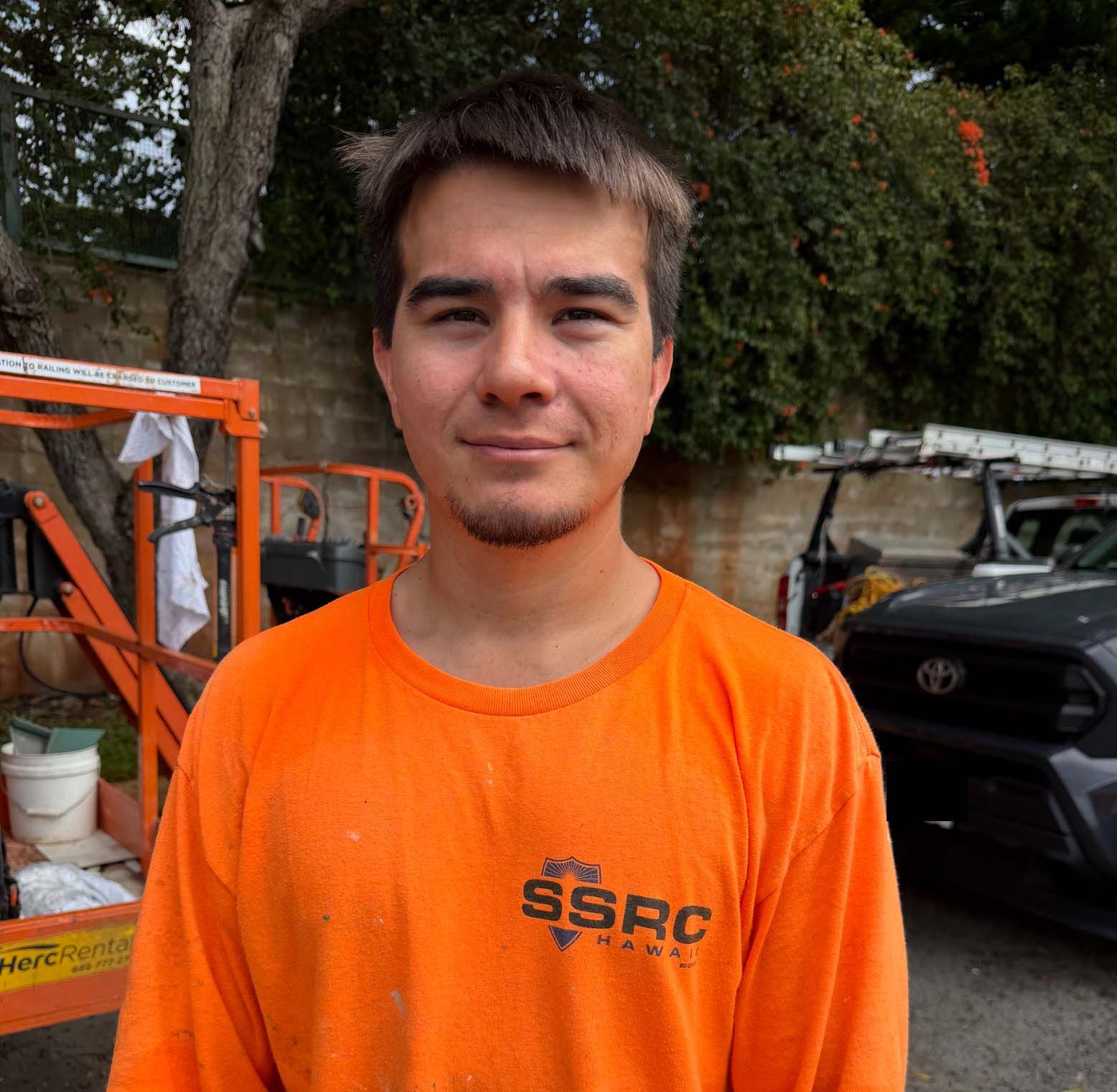 Person in an orange shirt standing outdoors near equipment and a parked car