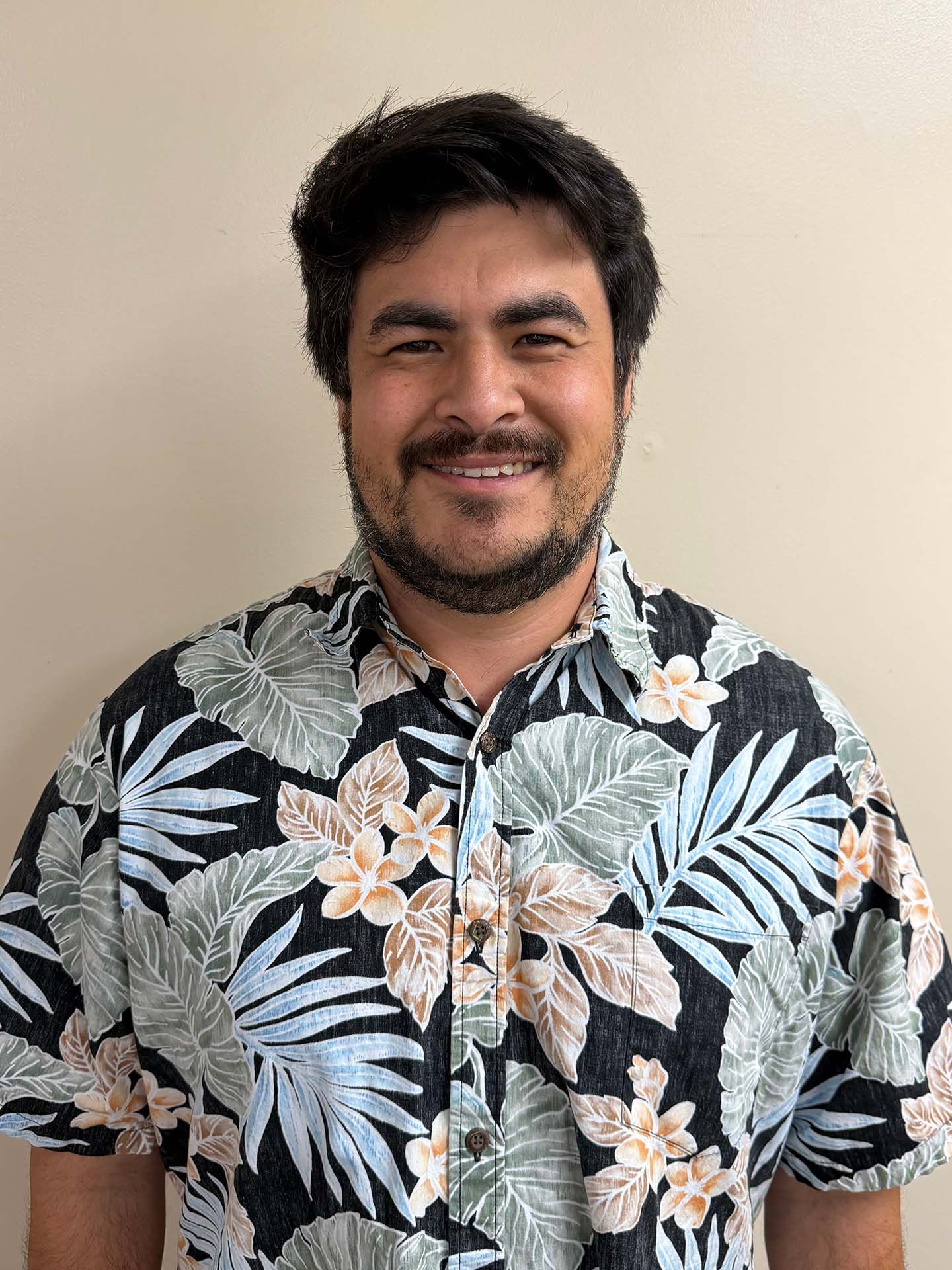 Smiling man in a black floral Hawaiian shirt against a plain beige wall