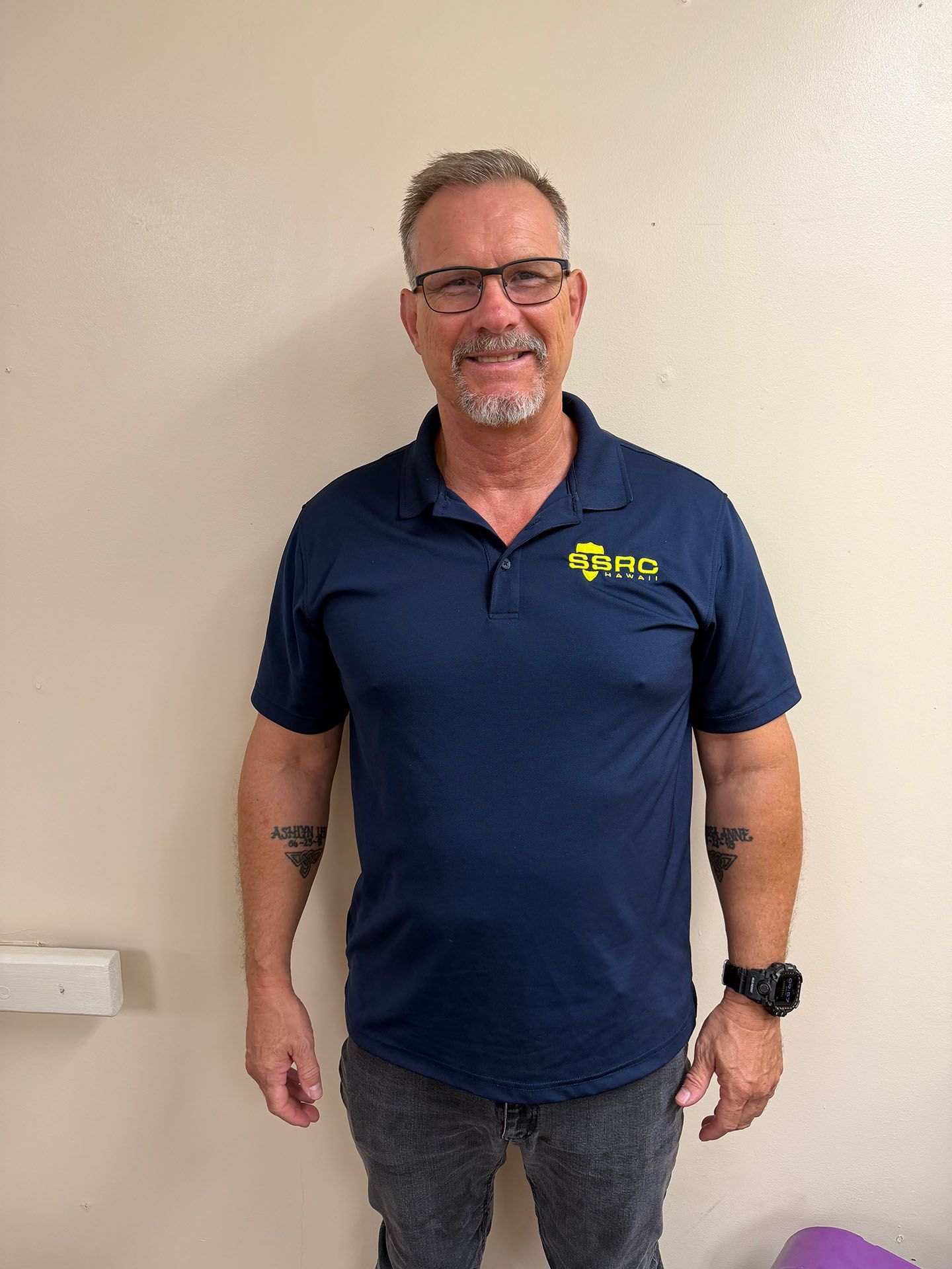 Man in navy polo shirt and gray jeans standing against a plain wall, smiling with arms at his sides
