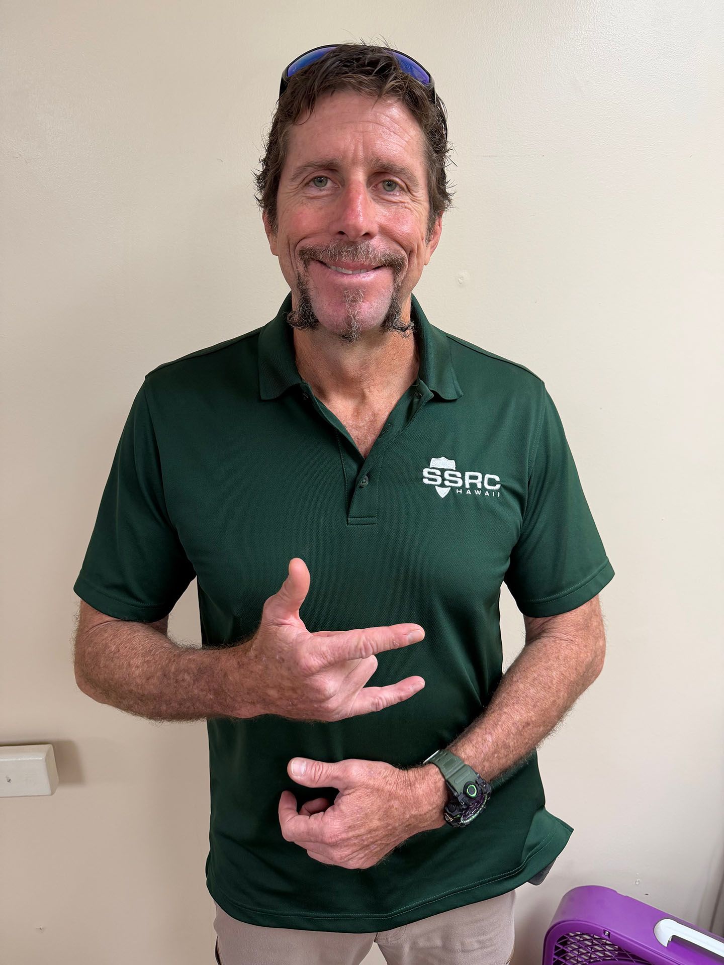 Man in a green polo smiling and making a hand sign against a plain wall