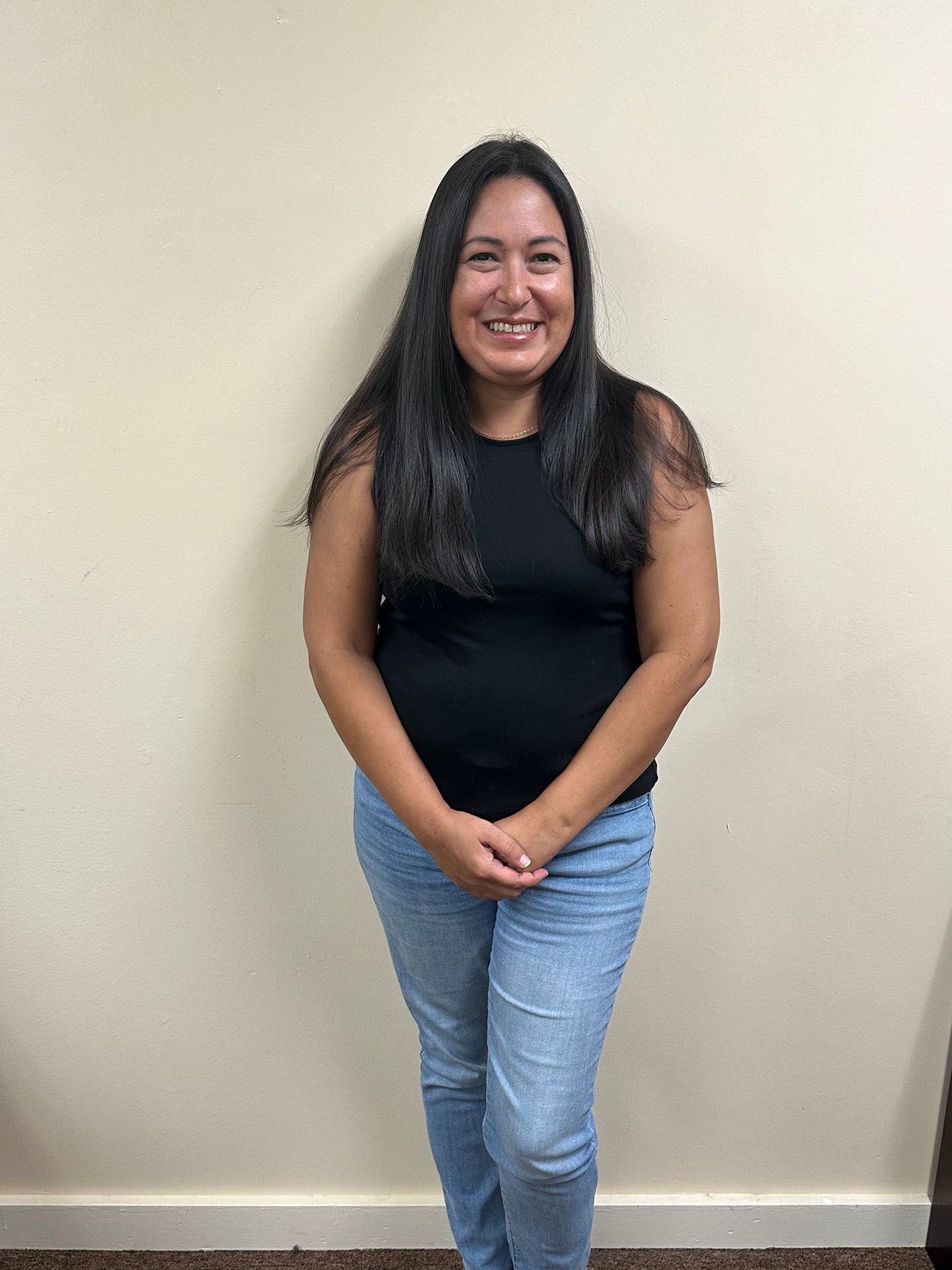 Person standing against a beige wall, smiling, wearing a black top and light blue jeans