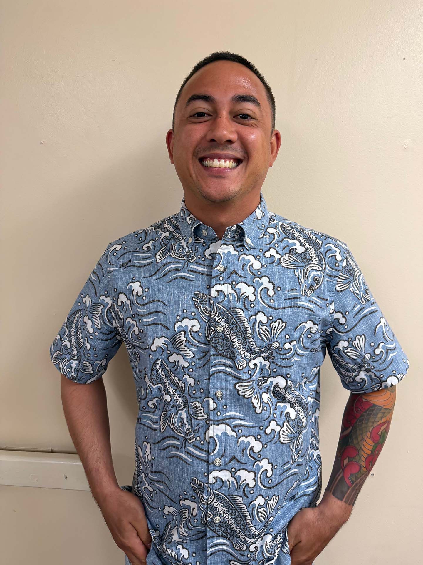 Smiling person in a blue floral shirt standing against a beige wall, hands on hips