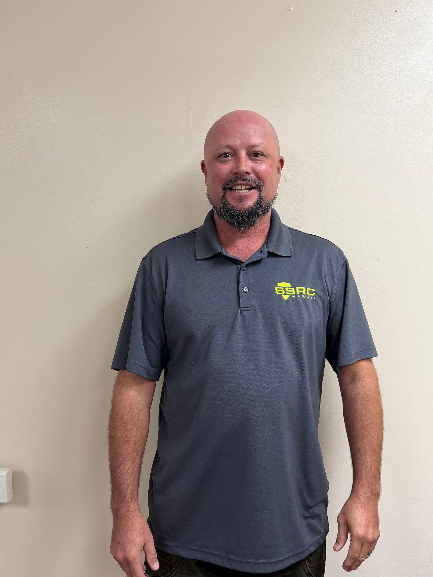 Smiling man in a gray polo shirt with a logo, standing against a plain wall