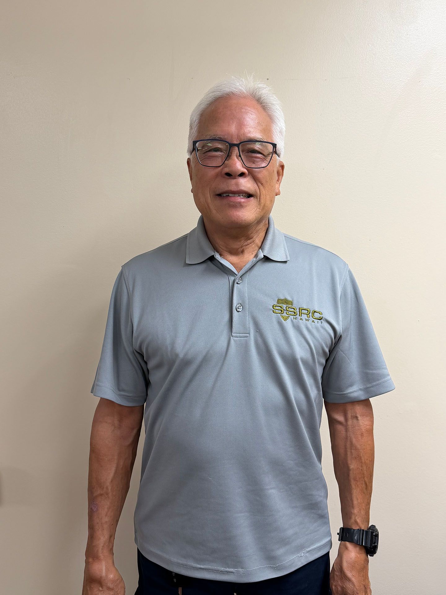 Man in a light gray polo shirt and glasses standing against a plain beige wall