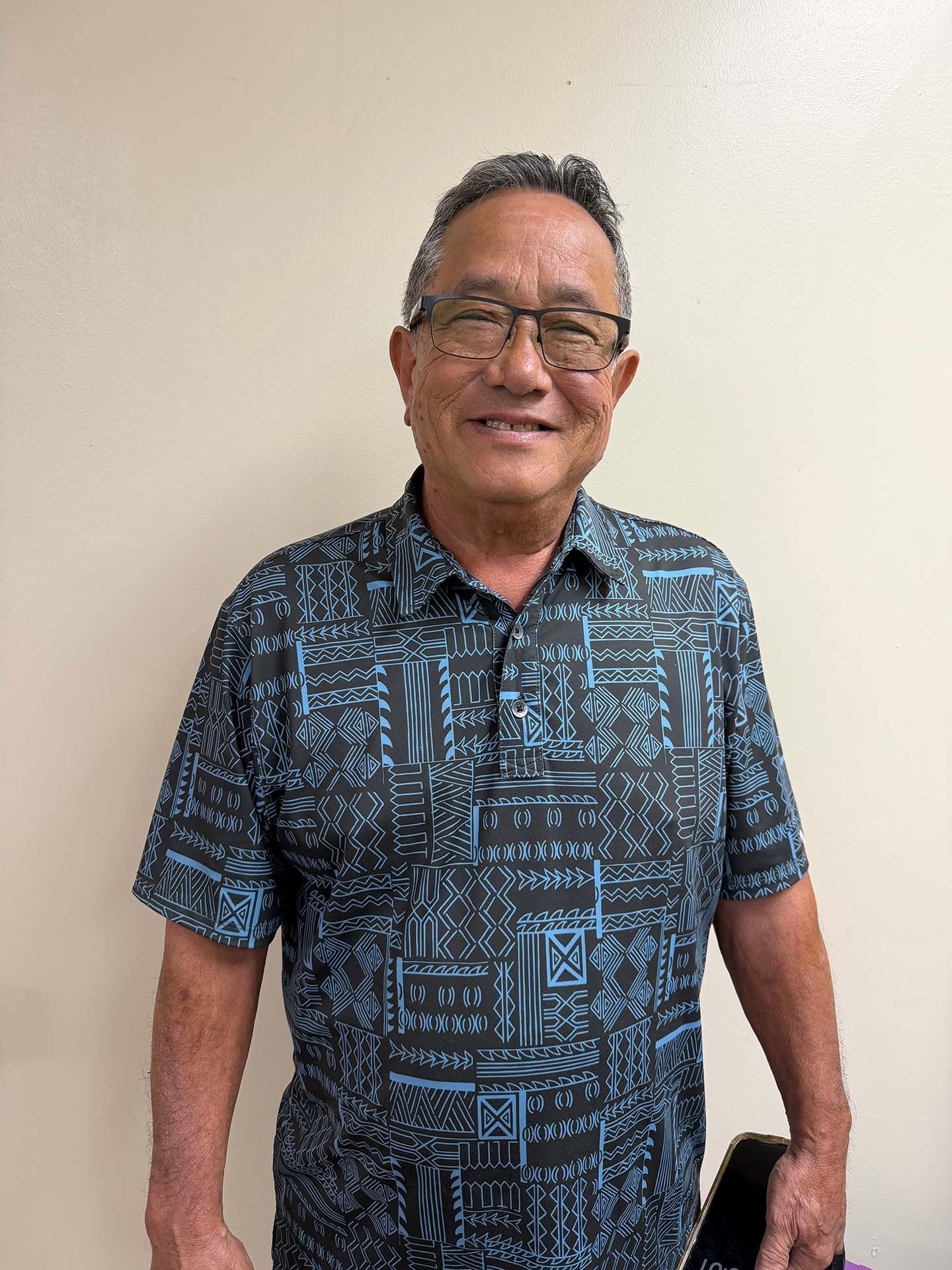 Smiling man in a blue patterned shirt holding a black phone against a plain wall