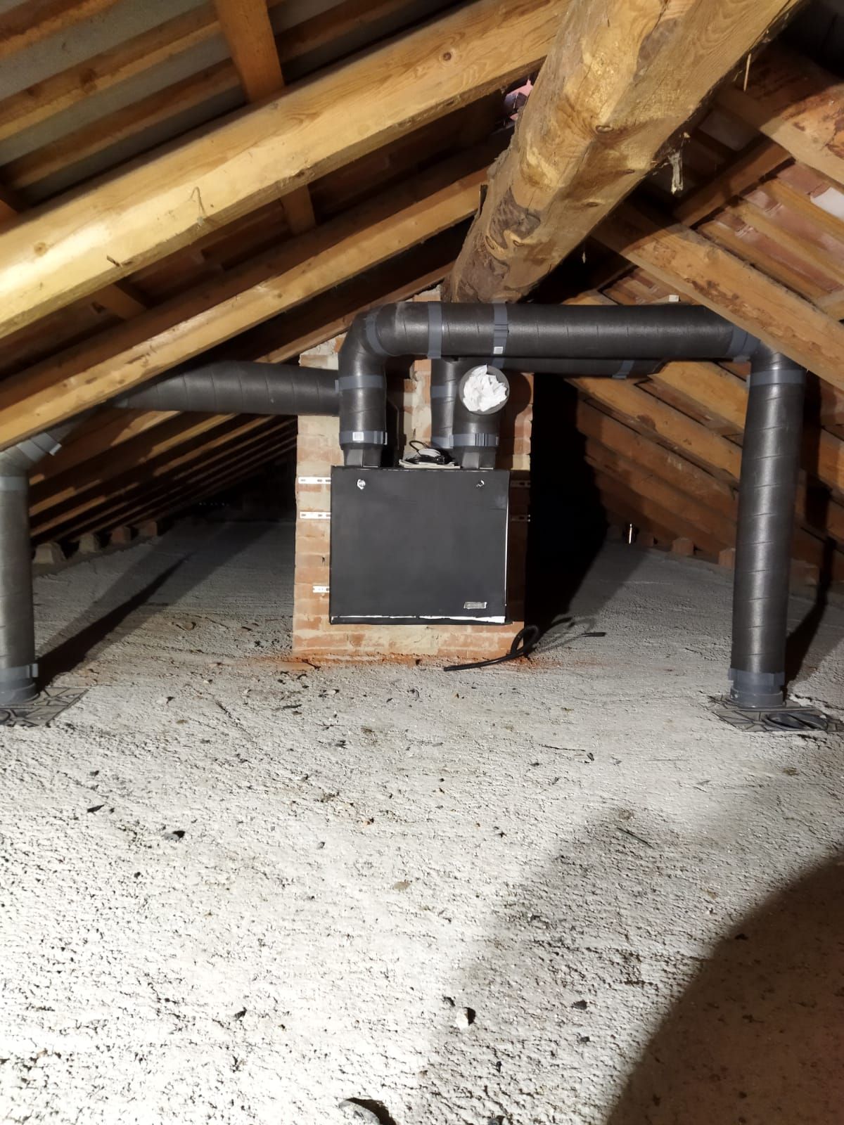Unità HVAC in soffitta, scatola nera, collegata a tubi grigi. Travi in legno sullo sfondo, isolamento grigio sul pavimento.