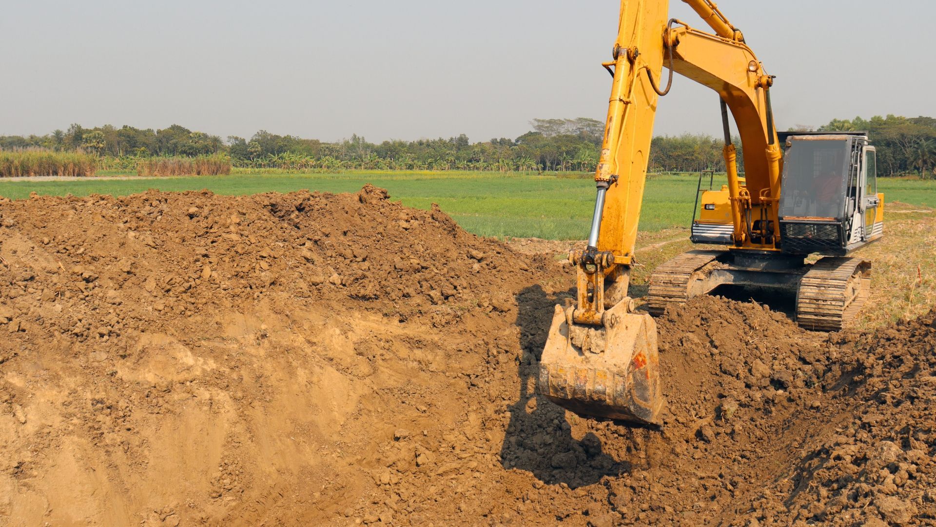 Yellow excavator digging earth in a field, with dirt pile and farmland in the background.