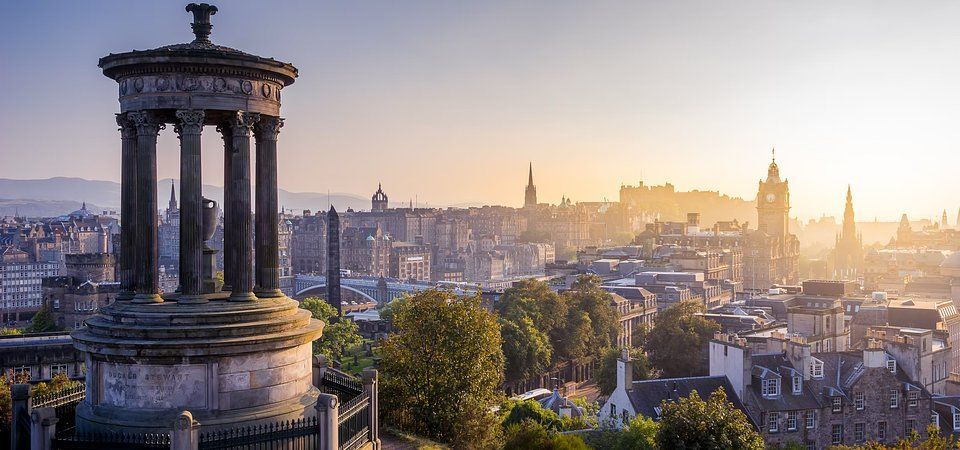Sunrise over Edinburgh skyline with a stone monument in the foreground and a cathedral spire in the distance