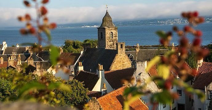 Stone church tower above rooftops and autumn leaves, with the sea in the background.