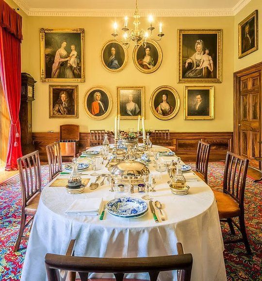 Formal dining room set for a meal, with a long table, white tablecloth, portraits, and chandeliers.