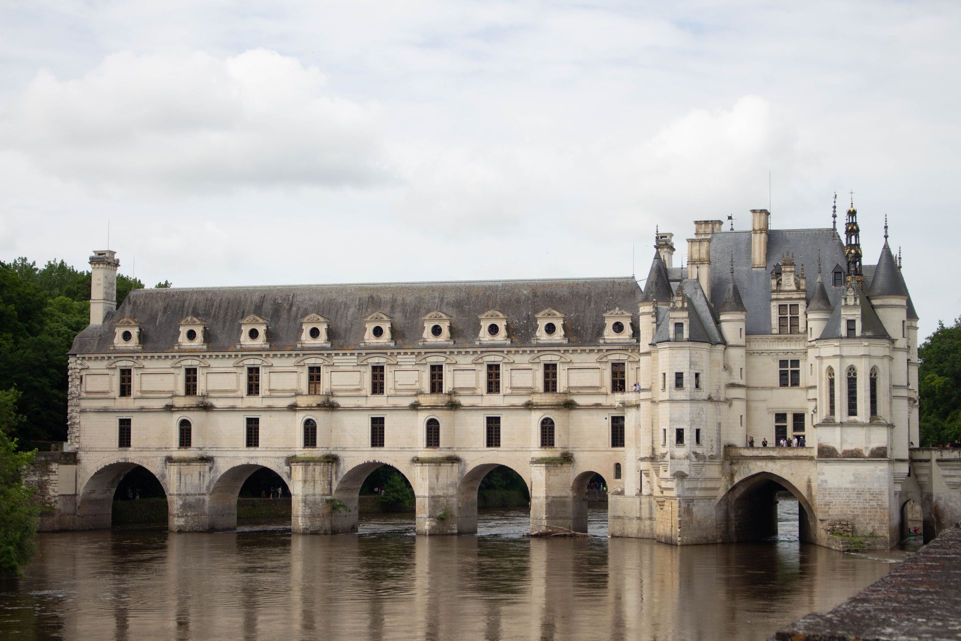Stone château with arched bridge over a river, reflected in calm water under a cloudy sky
