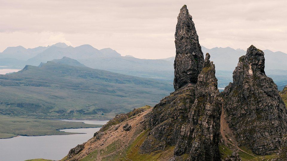 Jagged rock spires rise above a grassy ridge beside a lake, with misty mountains in the background.
