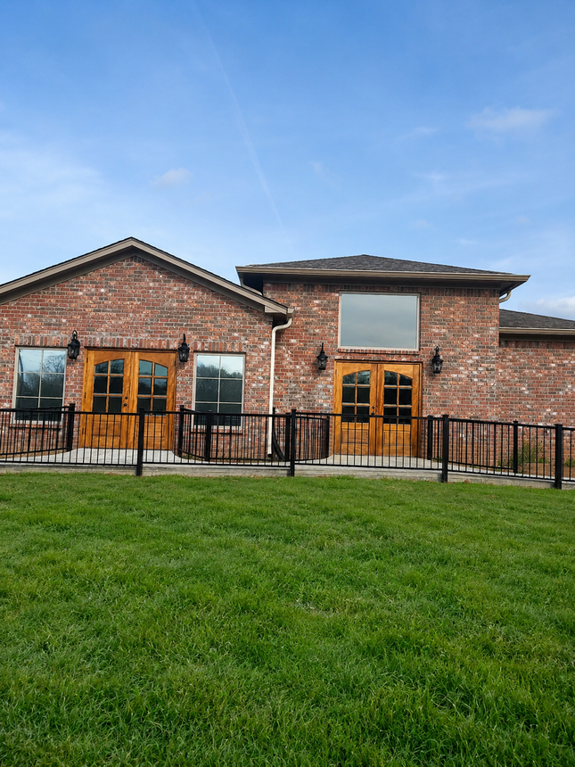 Brick house with wooden double doors, black metal fence, and dry yard under cloudy sky