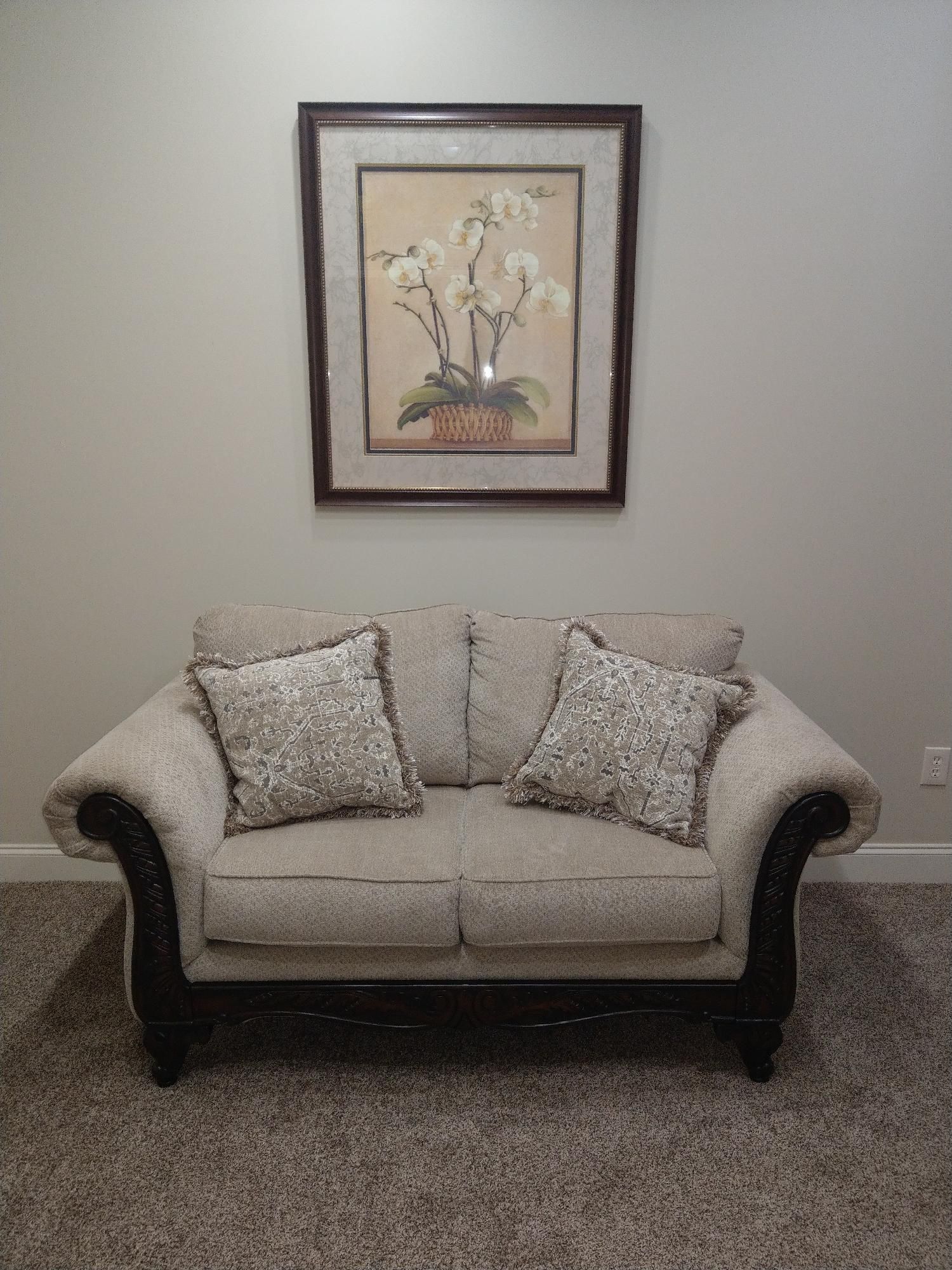 Beige loveseat with two patterned pillows beneath framed floral wall art on a neutral wall