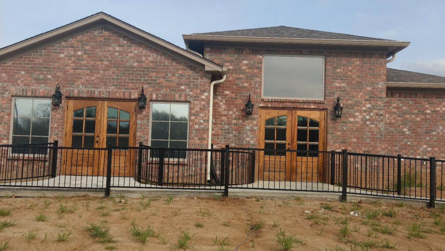 Brick house with wooden double doors, black metal fence, and dry yard under cloudy sky