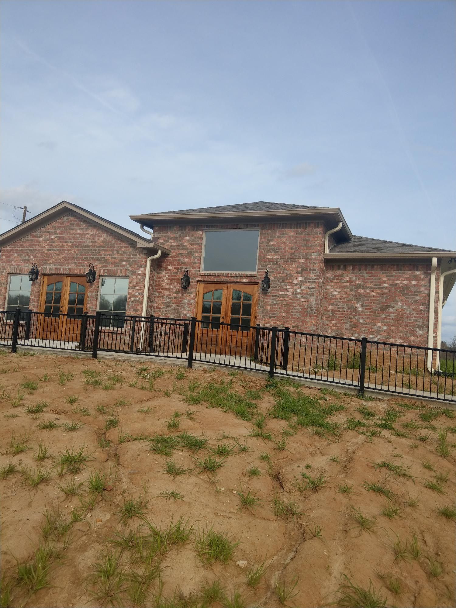 Brick house with wooden doors and black fence behind a sandy, grassy foreground under a cloudy sky