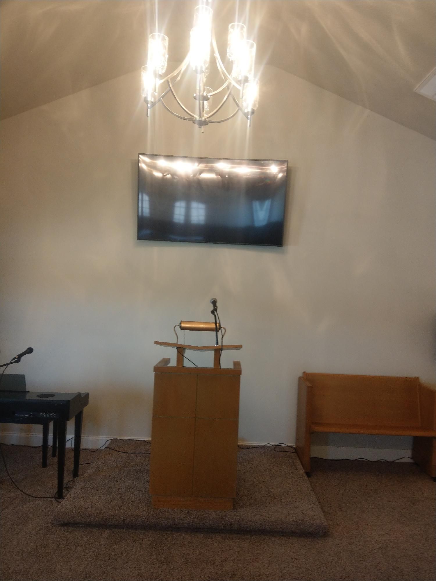 Small church altar with lectern, chandelier, wall-mounted screen, piano, and wooden bench on a carpeted platform.