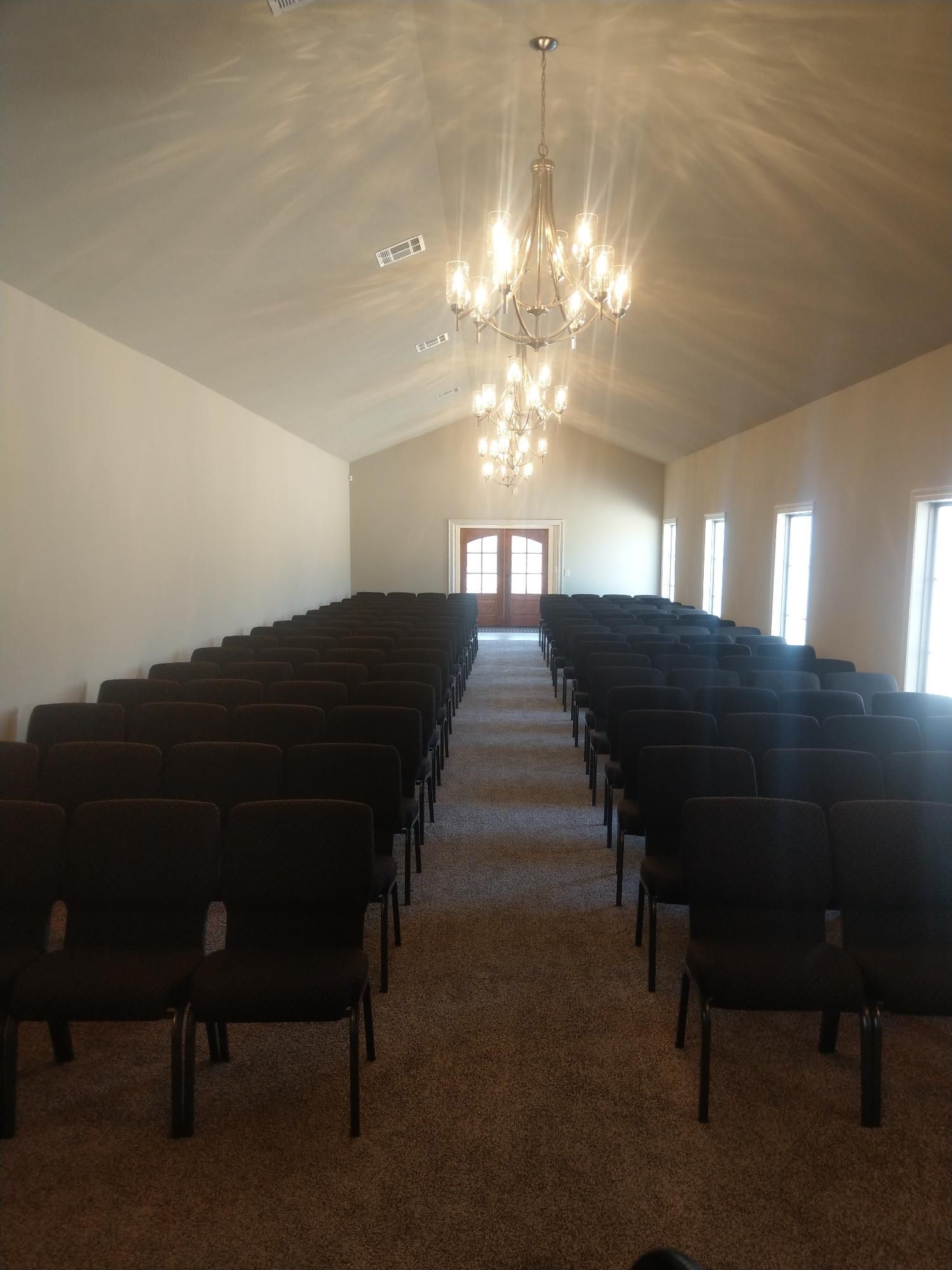 Long, narrow chapel or hall with rows of dark chairs facing a lit doorway and chandelier hanging overhead