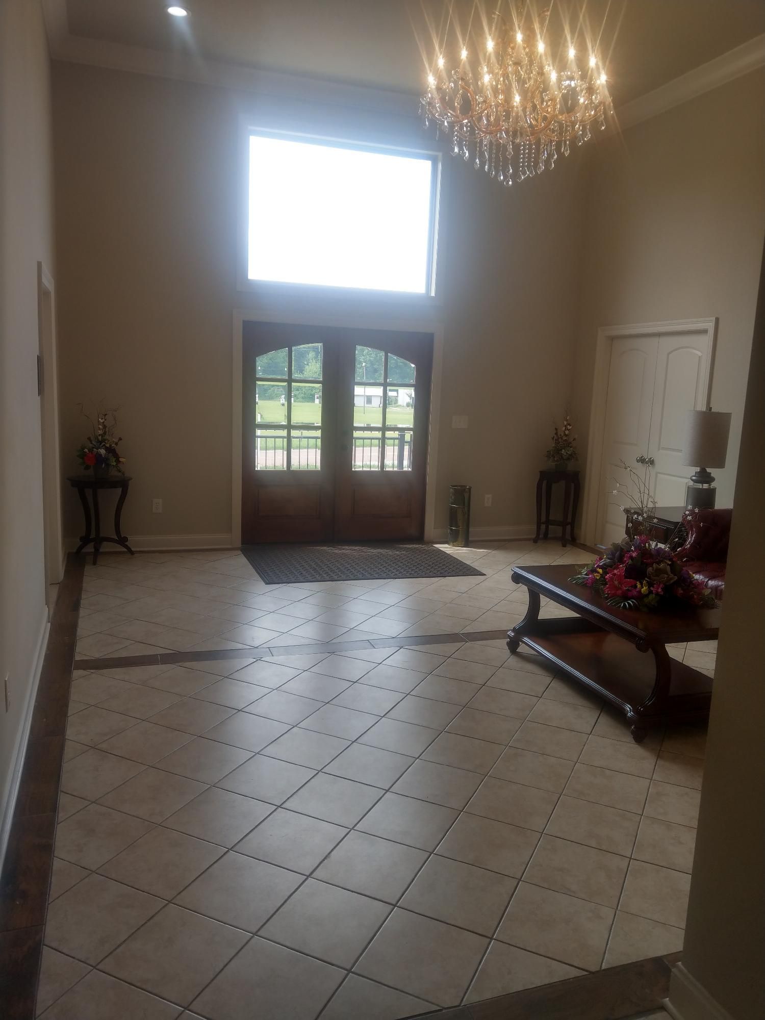 Bright foyer with tile floor, chandelier, double doors, and a bench with flowers.