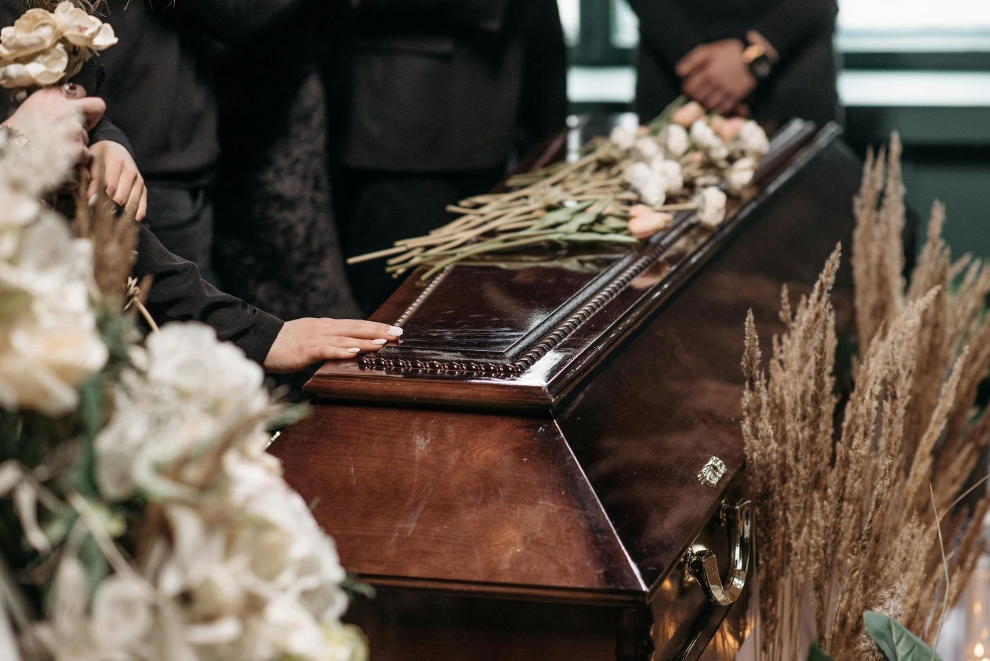 Wooden casket at a funeral, surrounded by white flowers and mourners in black attire