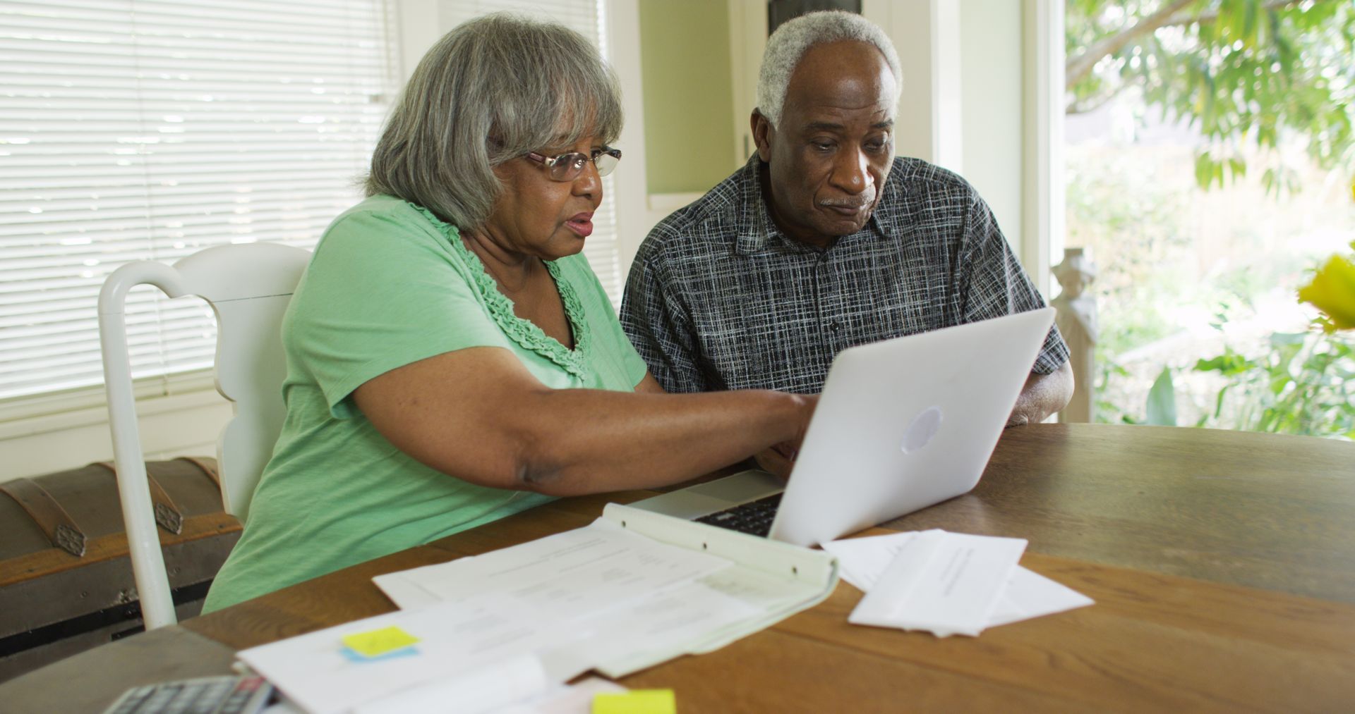 Two people sit at a table, looking at a laptop and papers in a bright room.