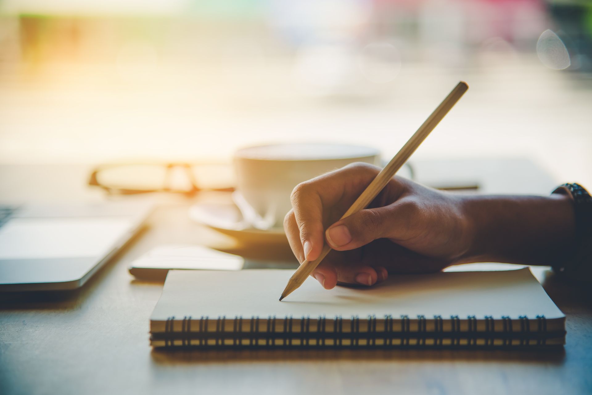 Hand writing in a spiral notebook beside a laptop and coffee on a sunlit desk