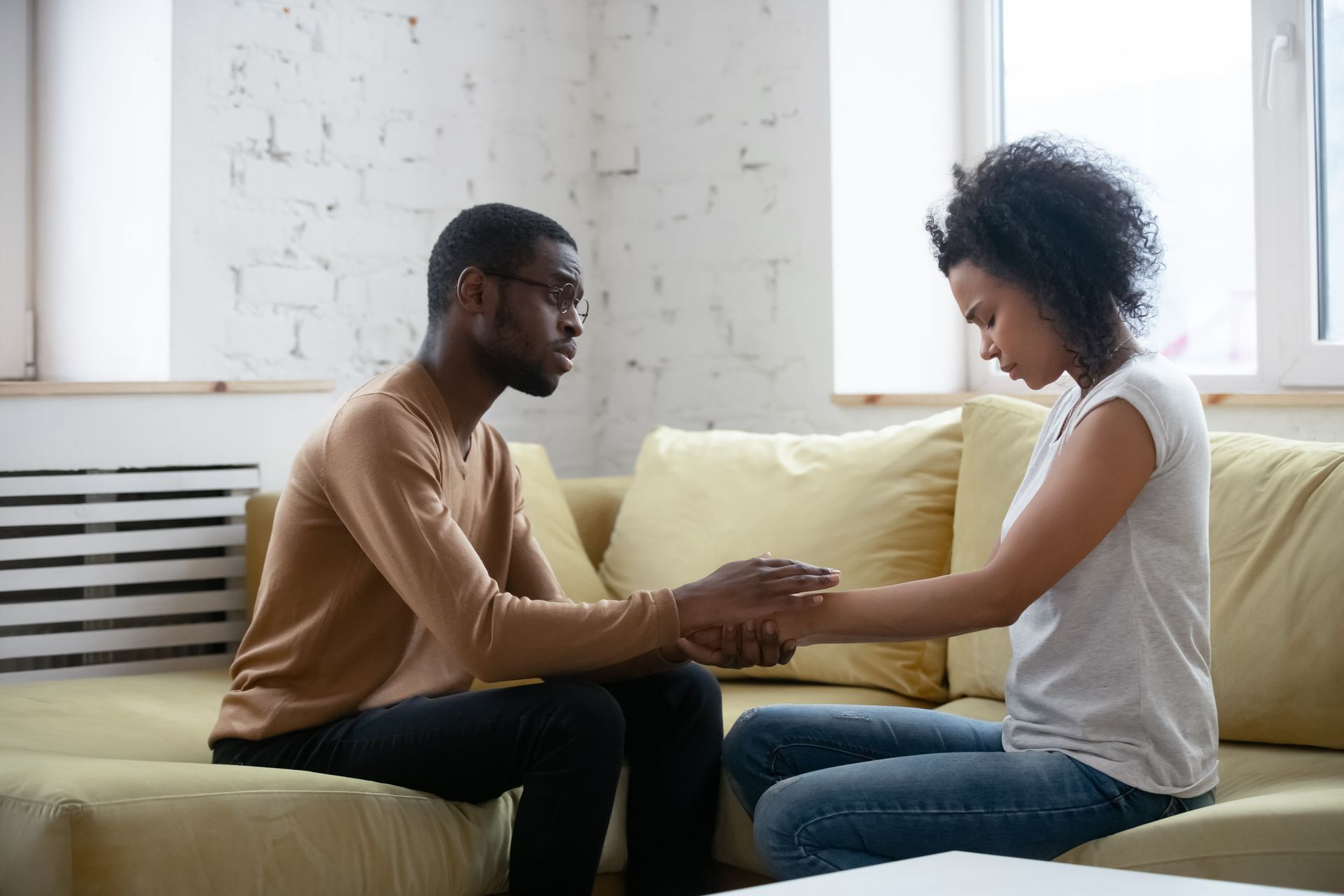 Two people sit facing each other on a couch, holding hands in a bright room.