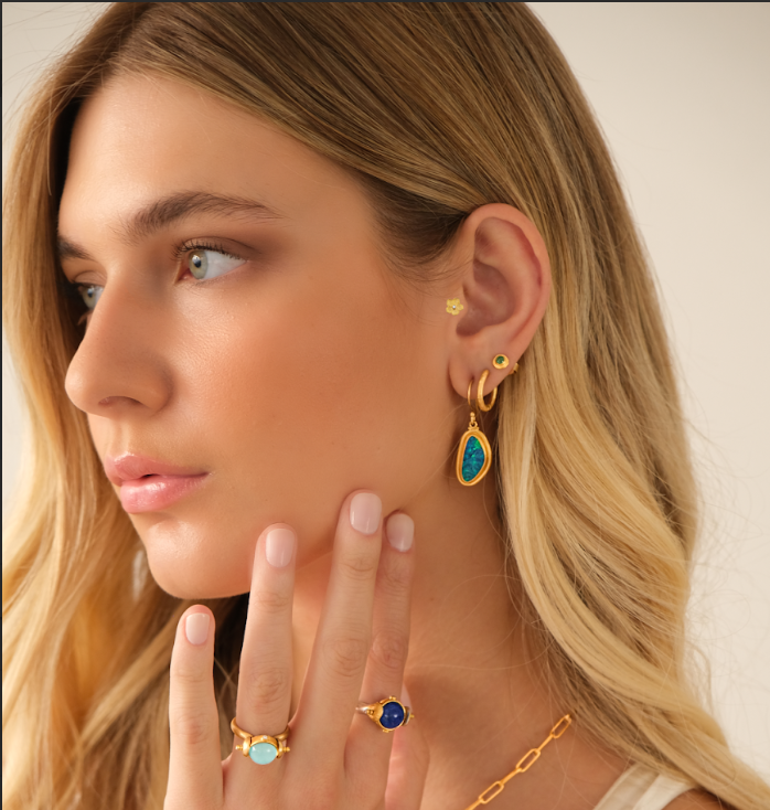 Woman wearing gold earrings and rings with blue and turquoise stones, touching her face.