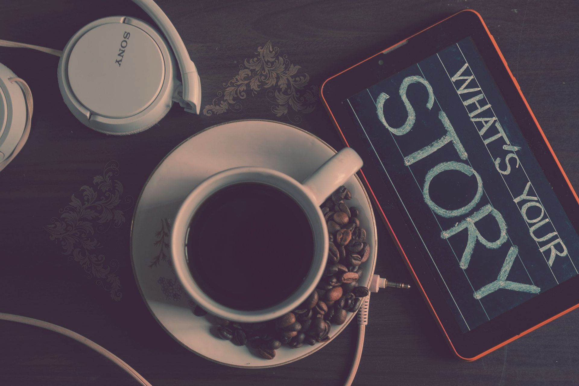 A cup of coffee surrounded by coffee beans, headphones, and a tablet displaying the words ‘What’s Your Story?’—a scene evoking creativity and reflection.
