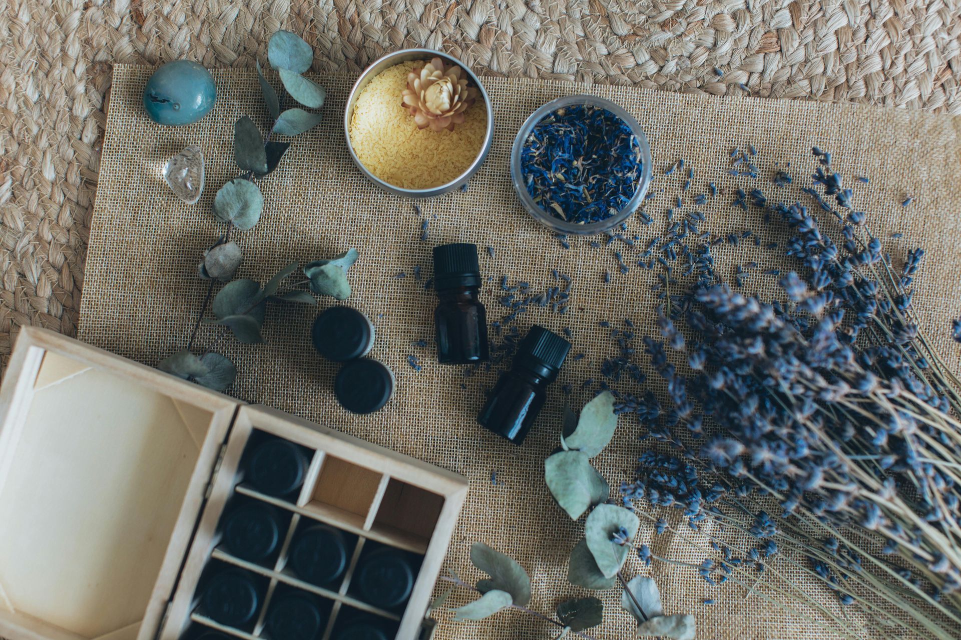 Top-down view of small essential oil bottles, dried lavender, and natural herbal ingredients on a woven mat.