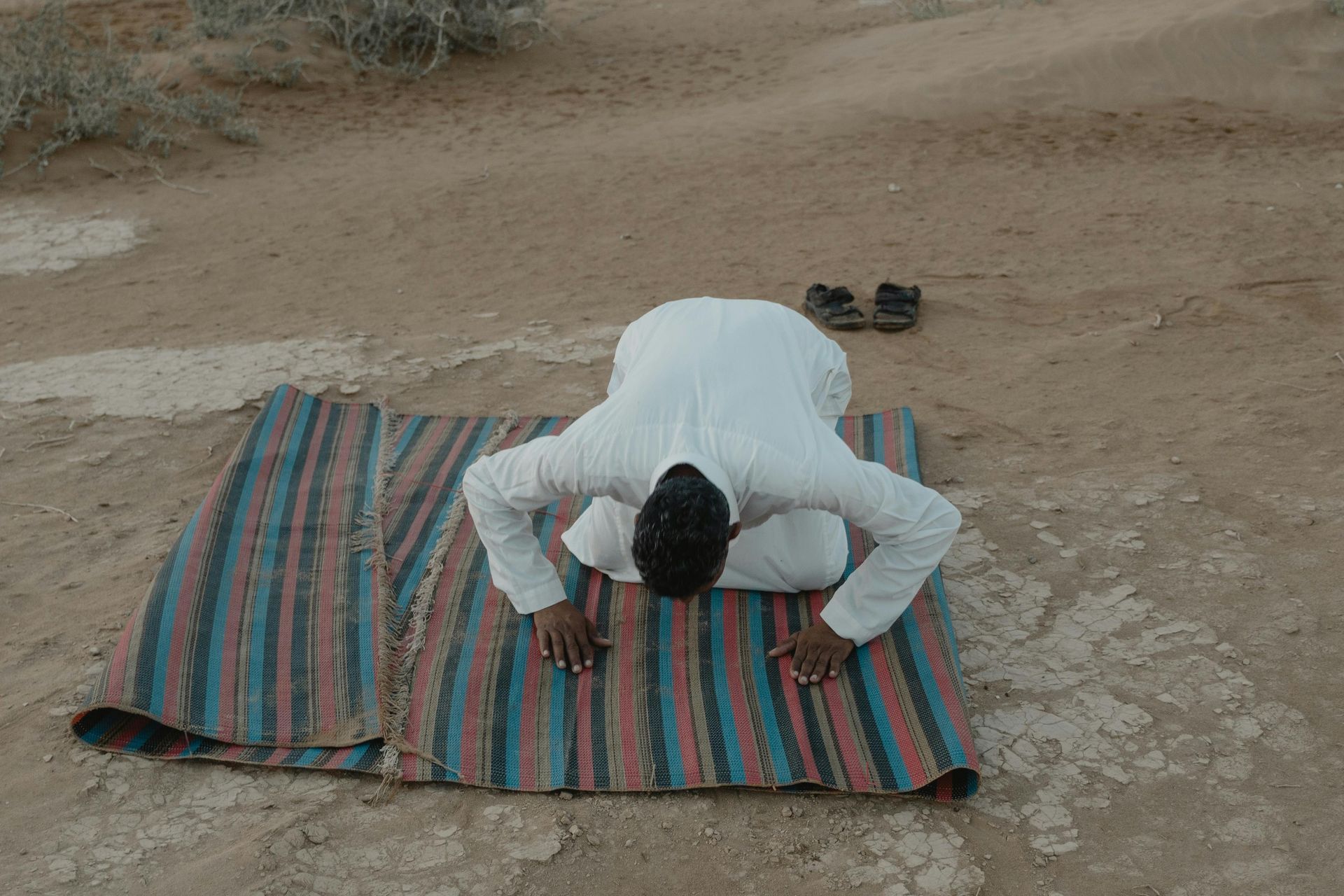 A man in a white robe bowing on a striped mat in a desert environment.