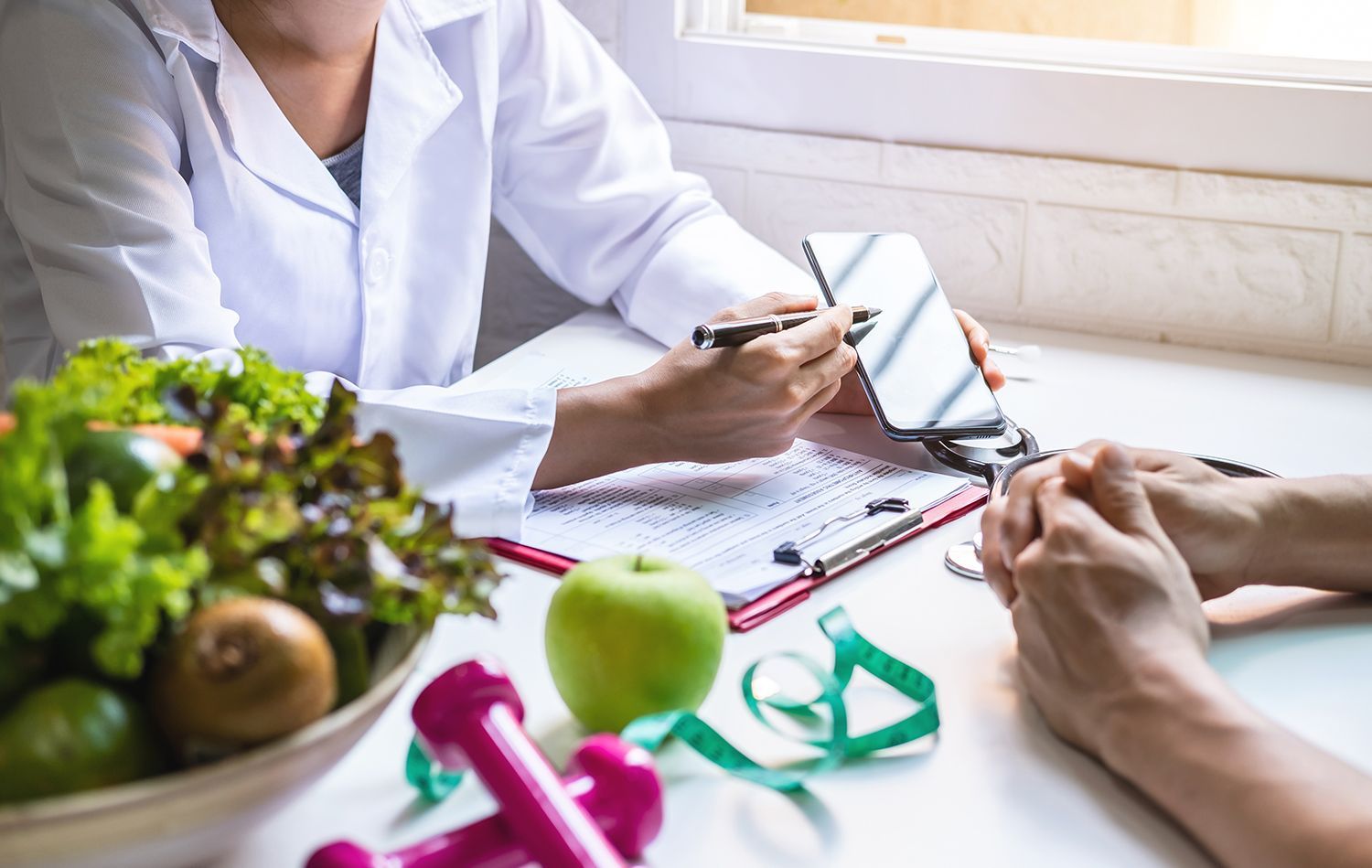 Dietitian pointing to a phone screen, discussing diet with a patient. Fresh produce and fitness equipment are on the table. Dietitian pointing to a phone screen, discussing diet with a patient. Fresh produce and fitness equipment are on the table.