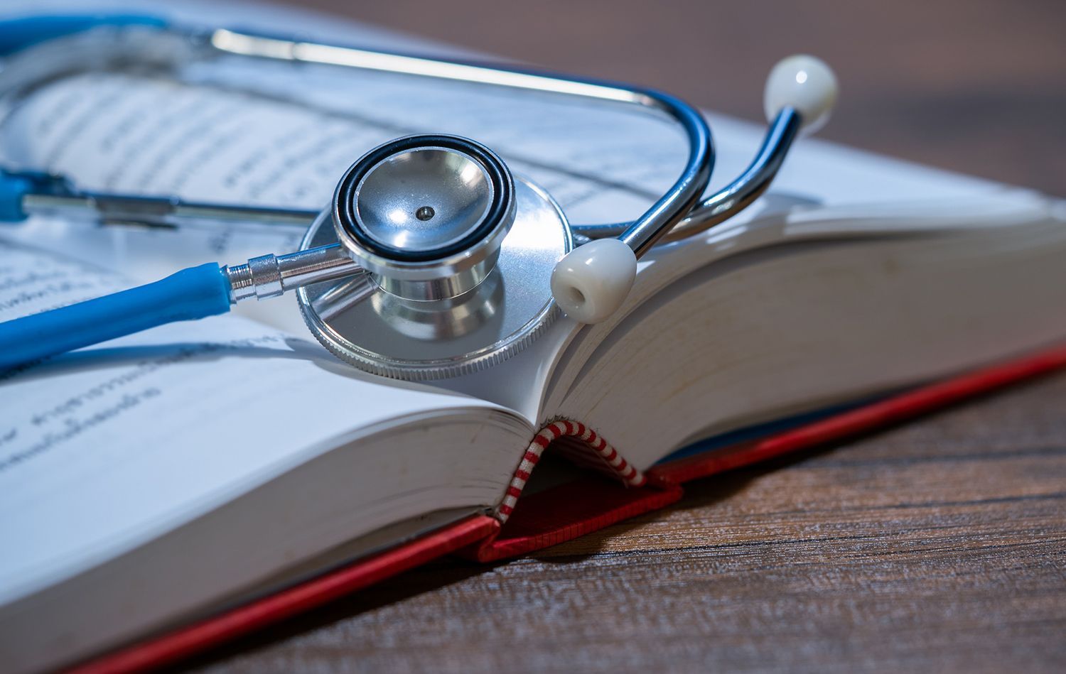 Blue stethoscope rests on an open book with red cover on wooden surface.