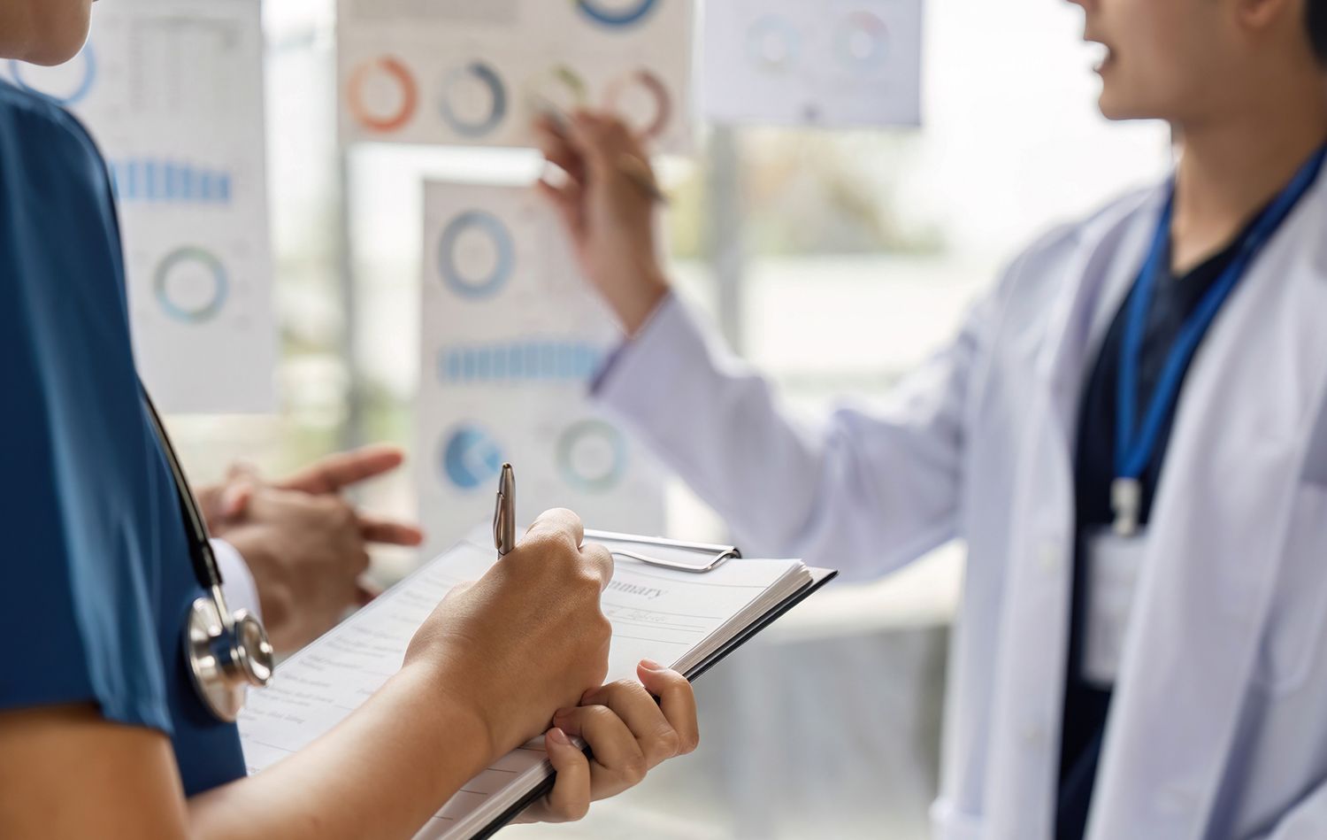 Two medical professionals analyzing charts, one writing on a clipboard, in a bright office.