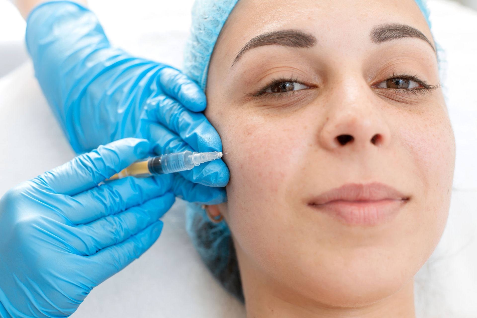 Woman receiving a facial injection in a medical setting, wearing a surgical cap.