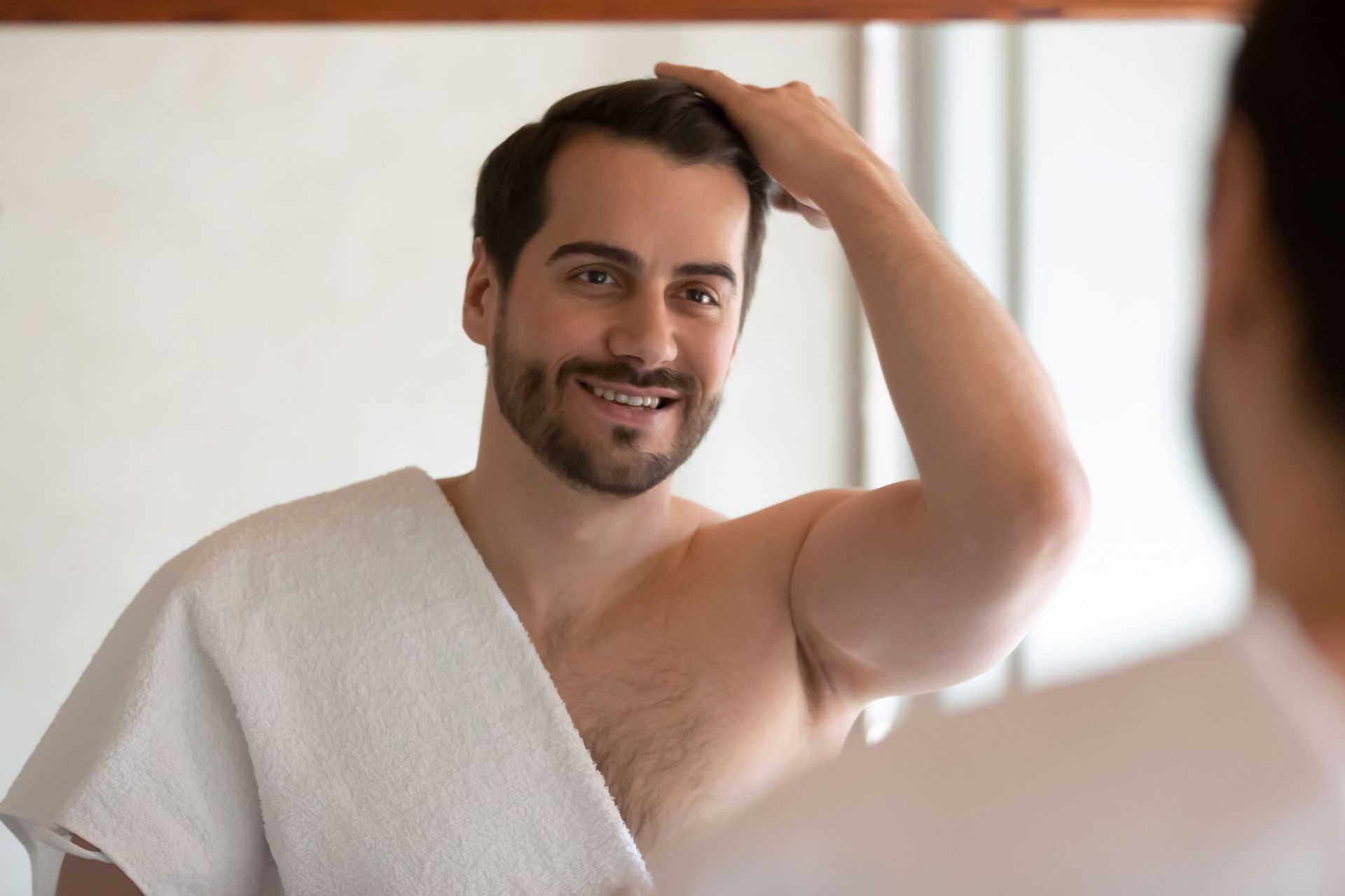 Man in a bathroom, looking in the mirror, smiling, and adjusting his hair with a towel draped over his shoulder. Man in a bathroom, looking in the mirror, smiling, and adjusting his hair with a towel draped over his shoulder.