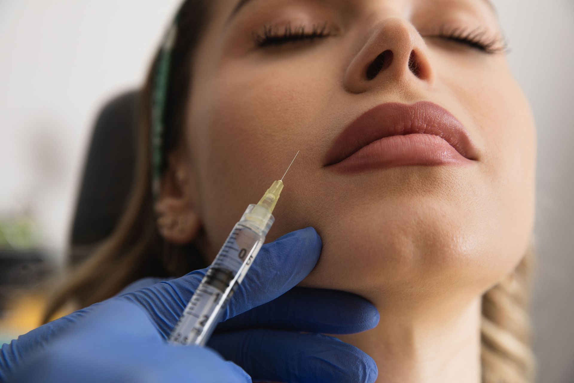 Person receiving a facial injection. A gloved hand holds a syringe near the chin of a person with eyes closed. Person receiving a facial injection. A gloved hand holds a syringe near the chin of a person with eyes closed.